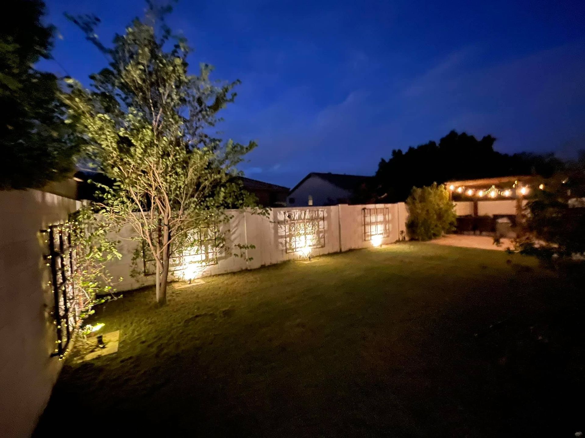 Backyard at dusk with lit fence, tree, lawn, and warm lights on a house patio