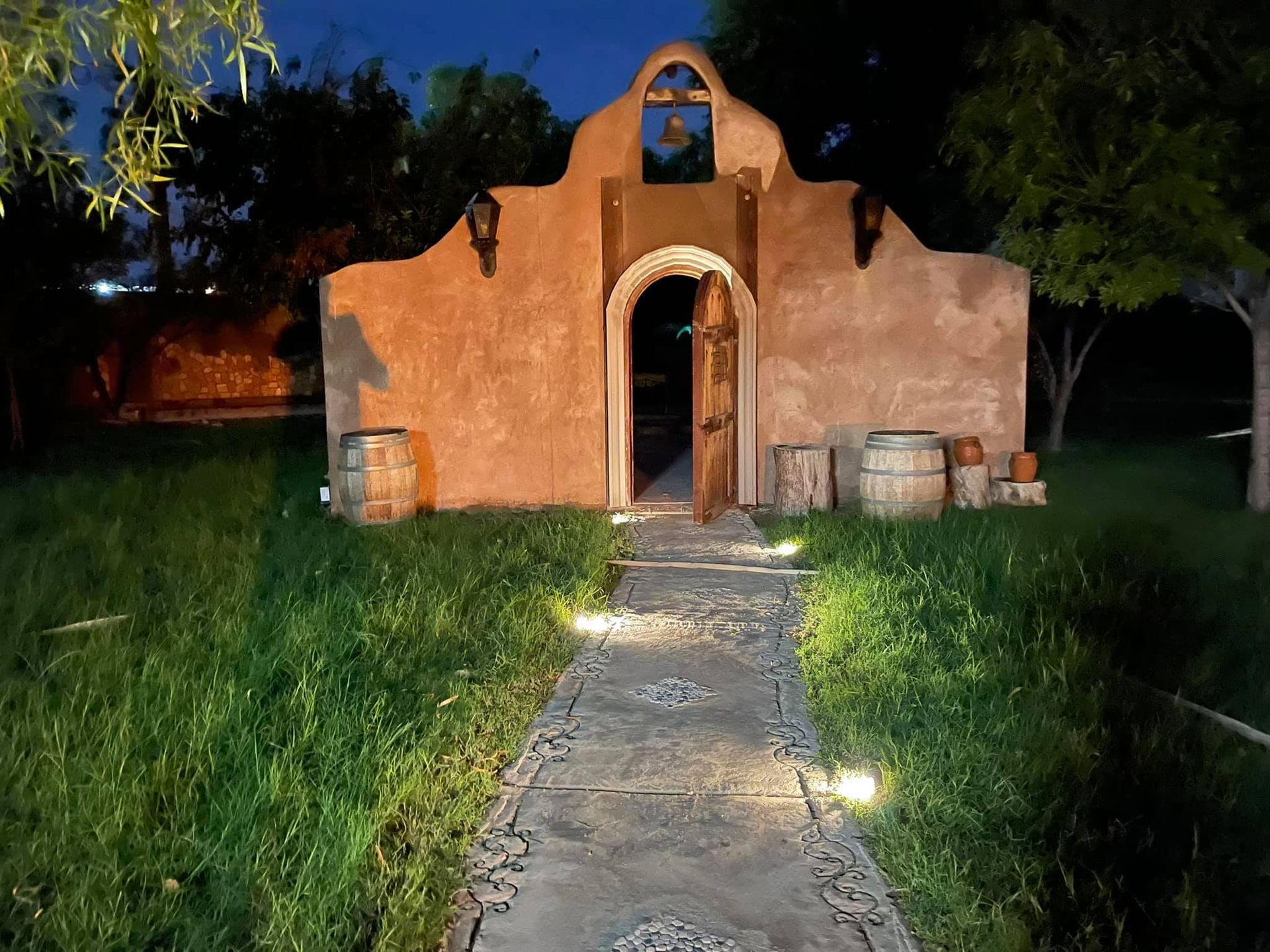 Small adobe-style chapel at night, lit path leading to open door, surrounded by grass and trees
