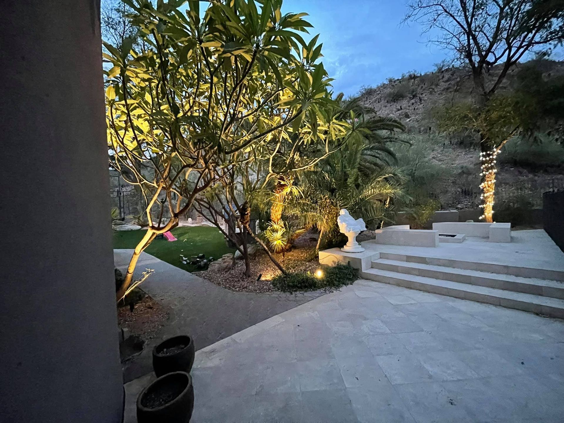 Lit patio at dusk with steps, tropical trees, and a hillside view