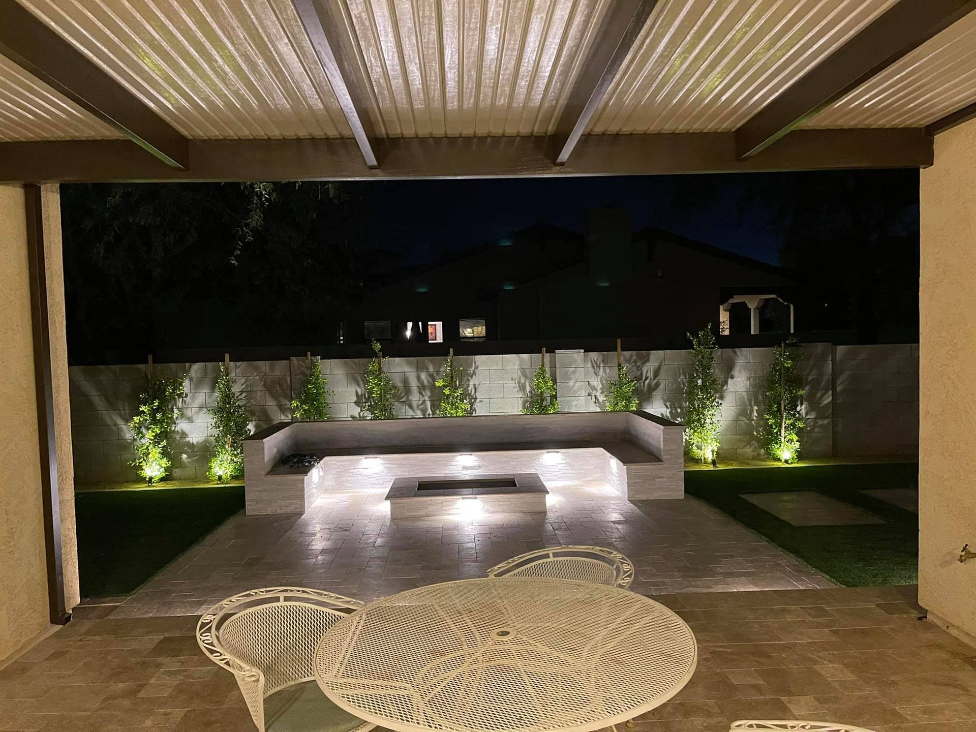 Modern patio at night with lit stone bench, water feature, and round woven table under a covered roof