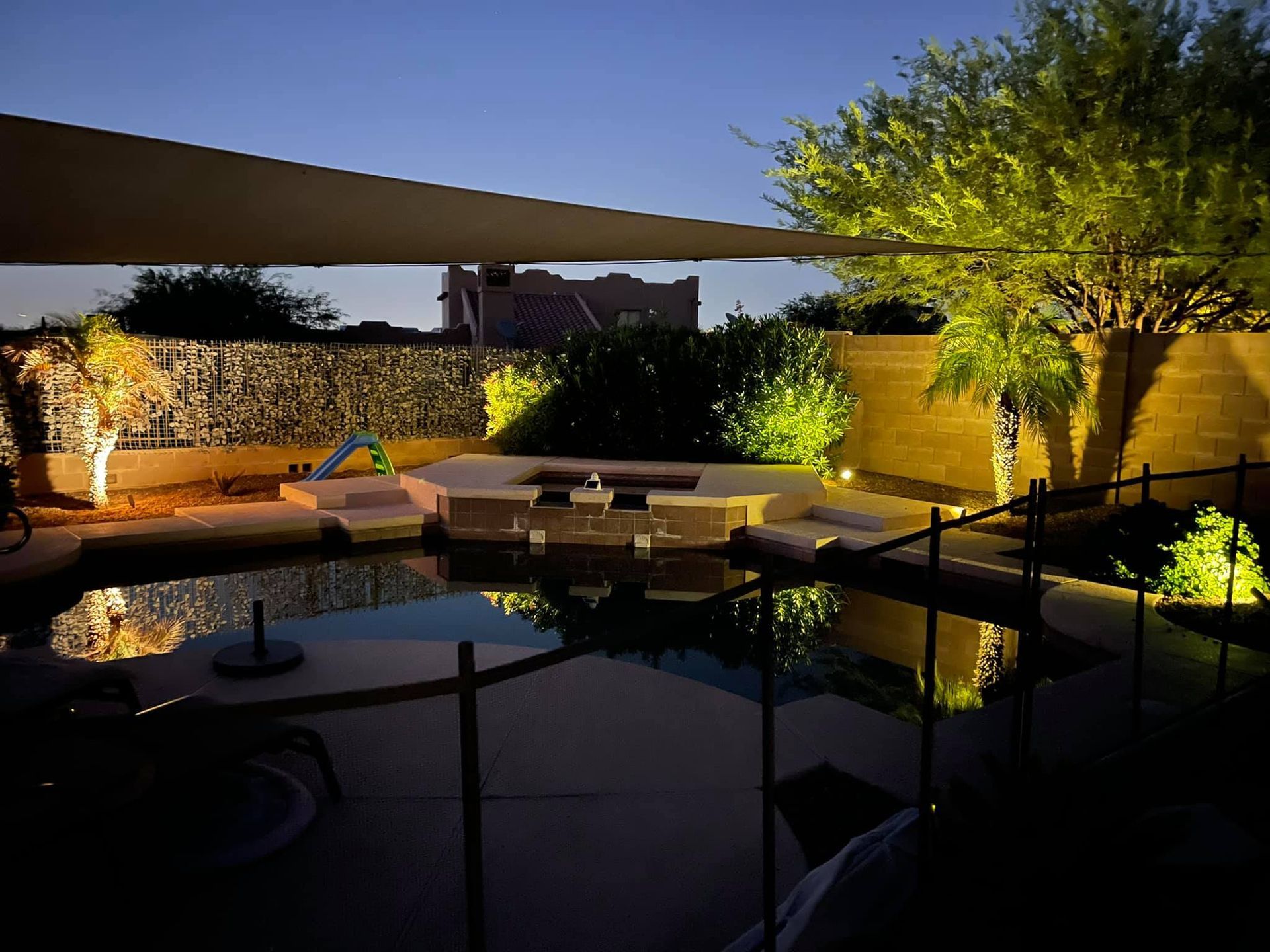 Modern backyard patio at dusk with a shaded seating area, stone wall, and lit trees beside a pool