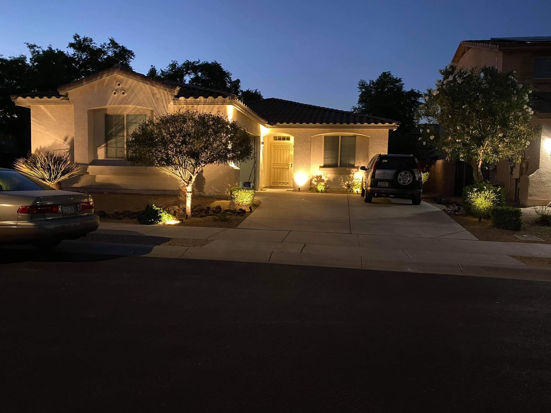 Well-lit suburban house at dusk with cars in the driveway and porch lights on