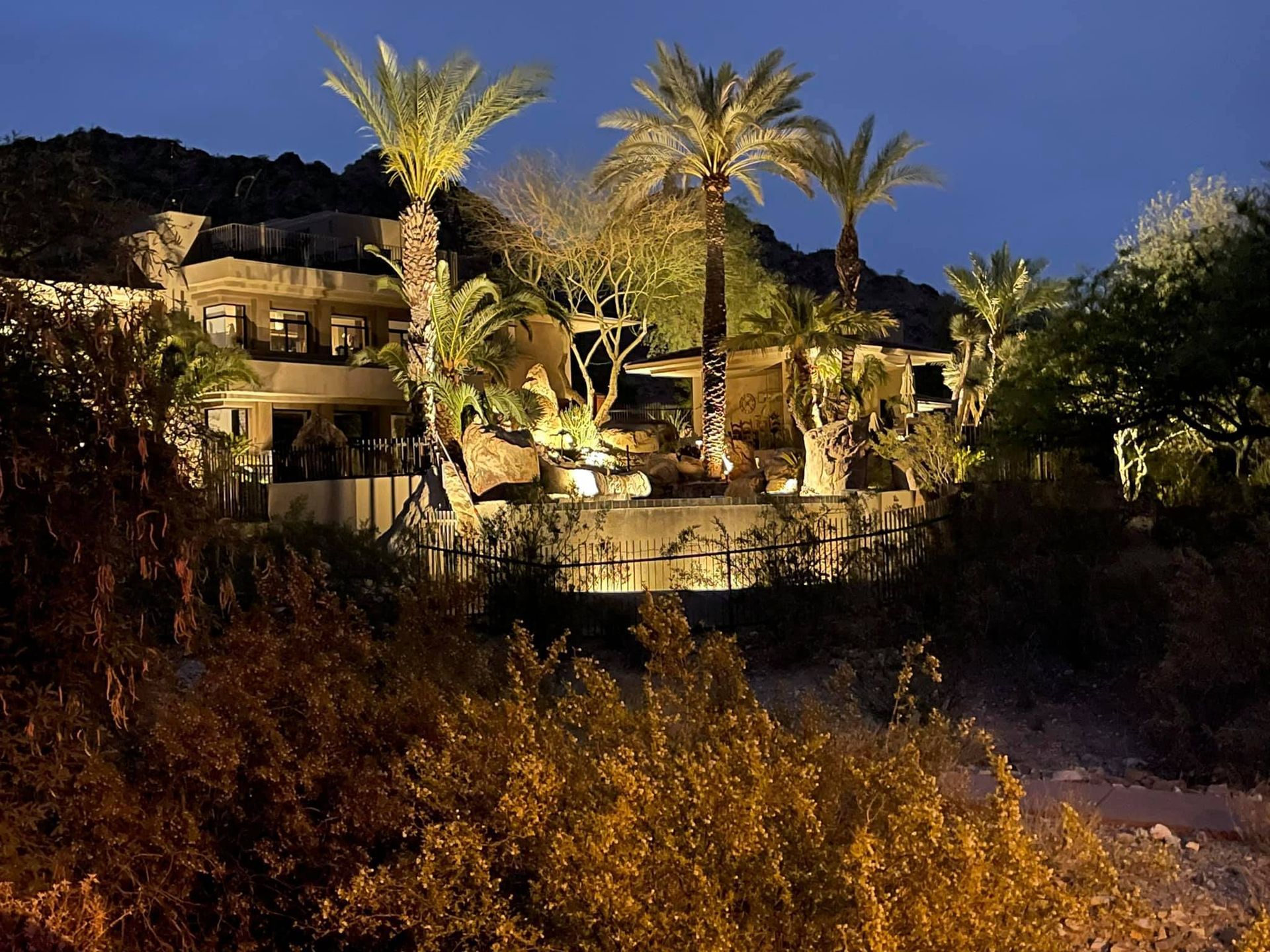 Illuminated hillside resort with palm trees at dusk, overlooking dark foliage and a lit pool terrace