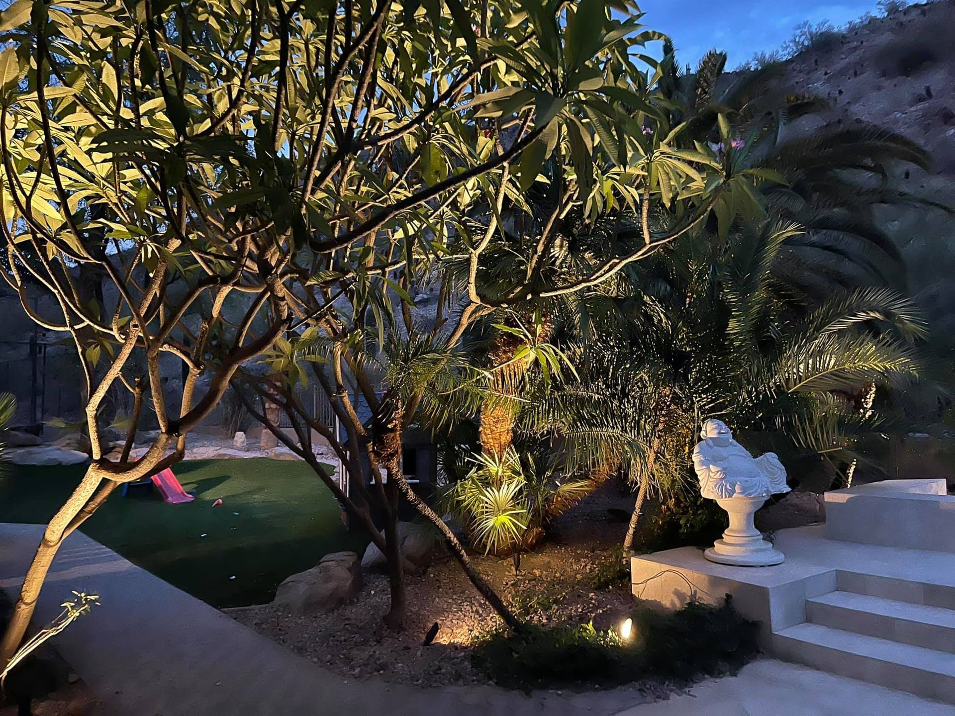 Dimly lit garden path with trees, palm plants, and white steps beside a decorative statue at dusk