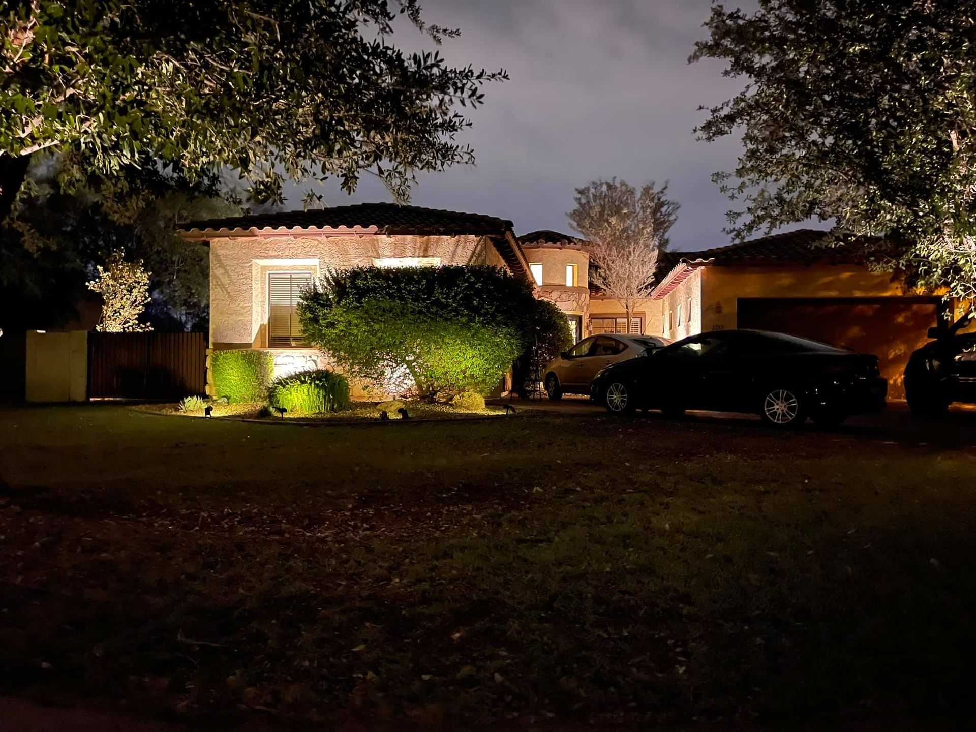 Nighttime view of a house with lit windows, bushes, and a parked car in the driveway