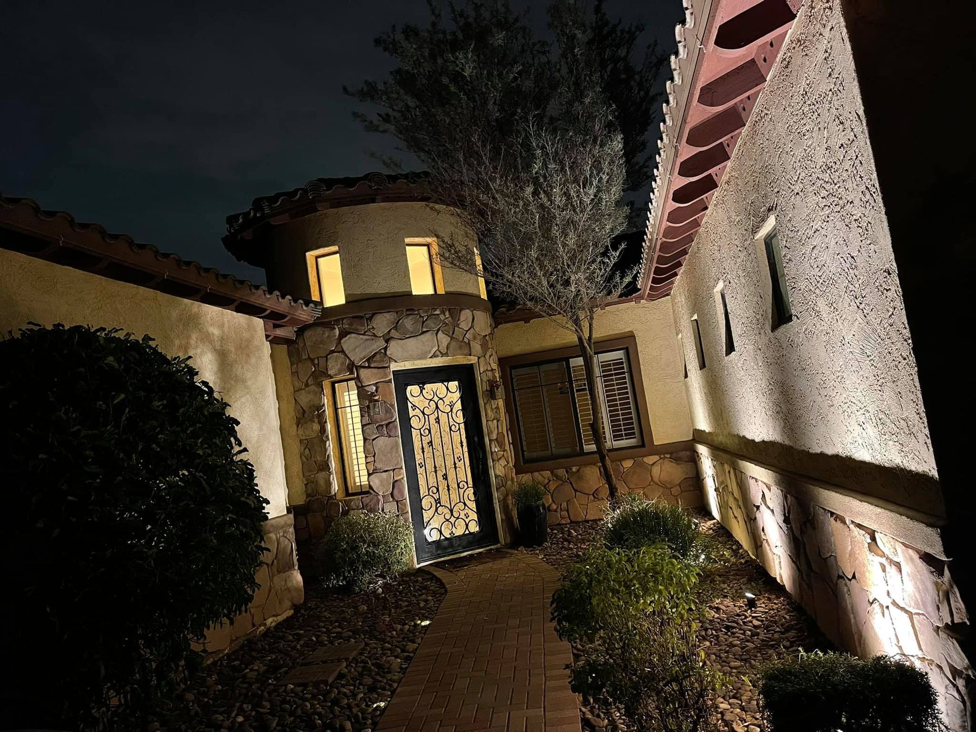 Nighttime view of a stucco house entry path with lit windows and outdoor wall lights.