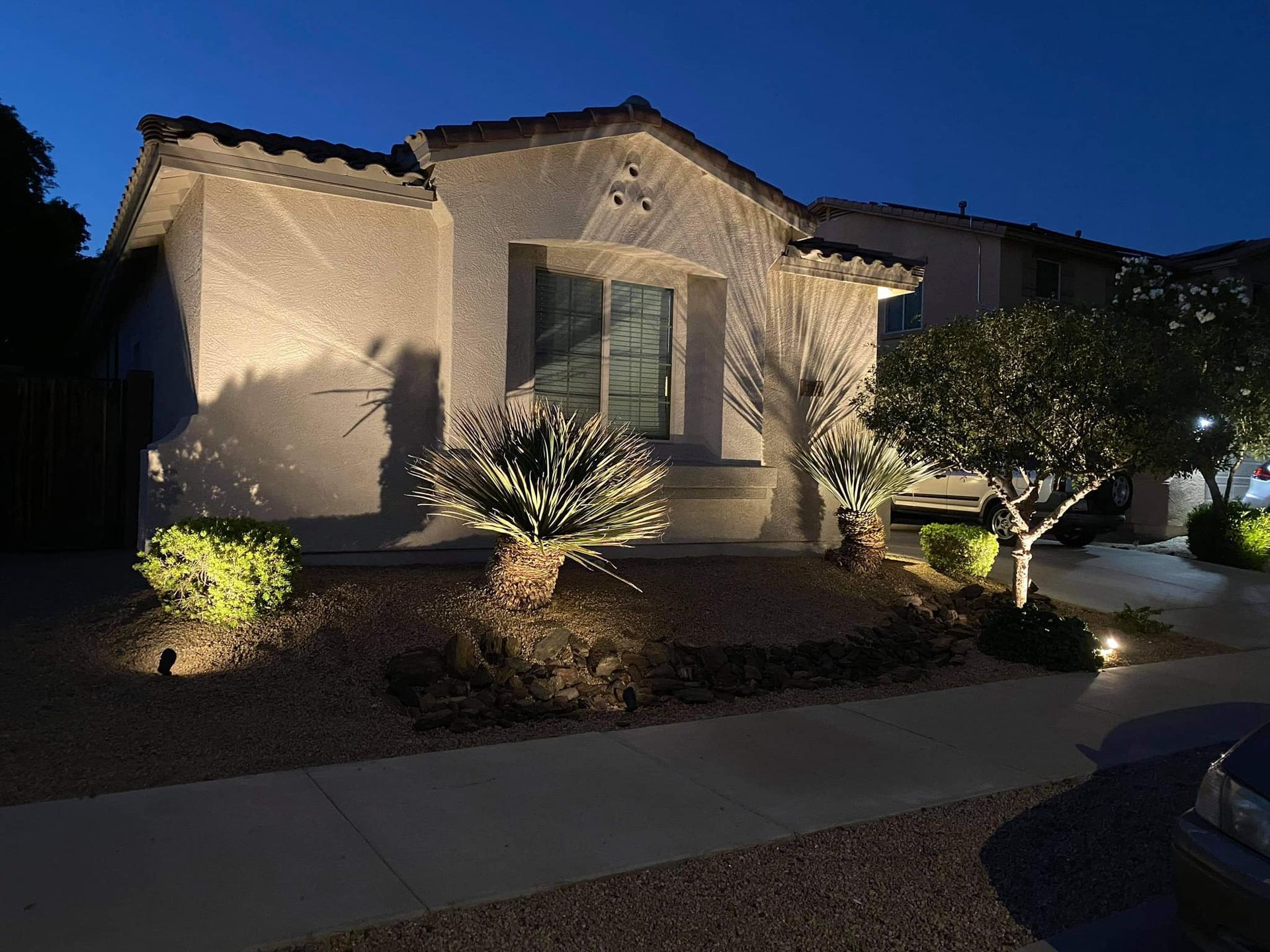 Single-story stucco house at dusk with lit landscaping and a sidewalk in front