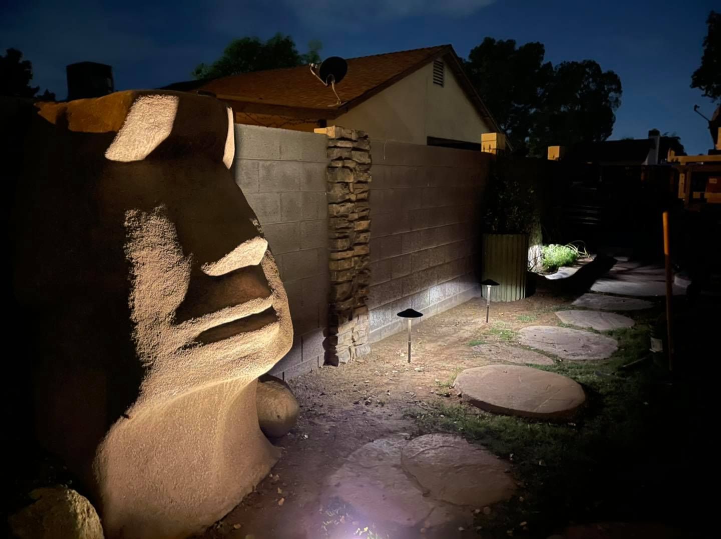 Dimly lit backyard with a lit stone sculpture, patio lights, and stepping stones at dusk