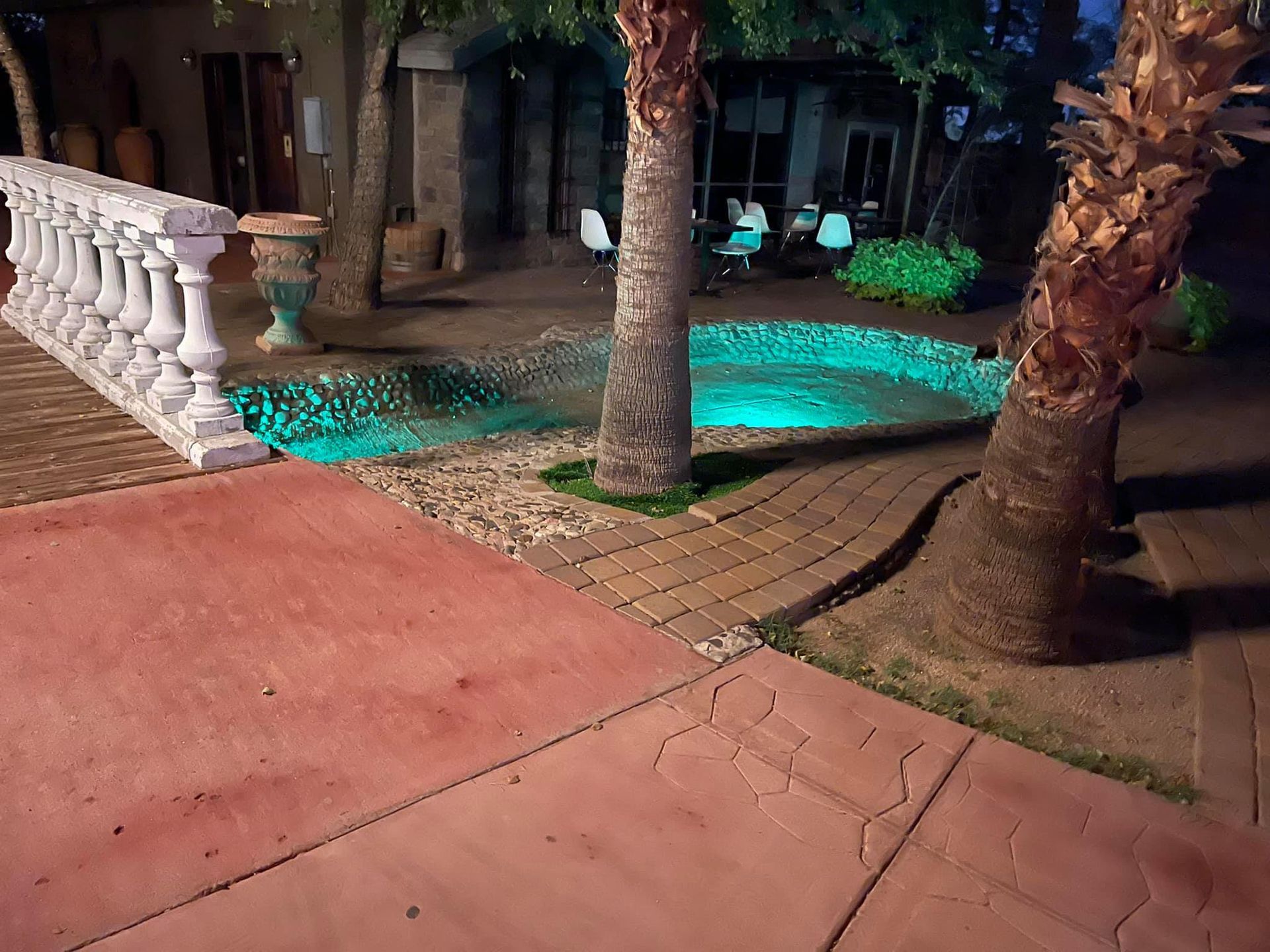 Patio with red pavement, palm trees, white railing, and turquoise-lit pool beside a building at night