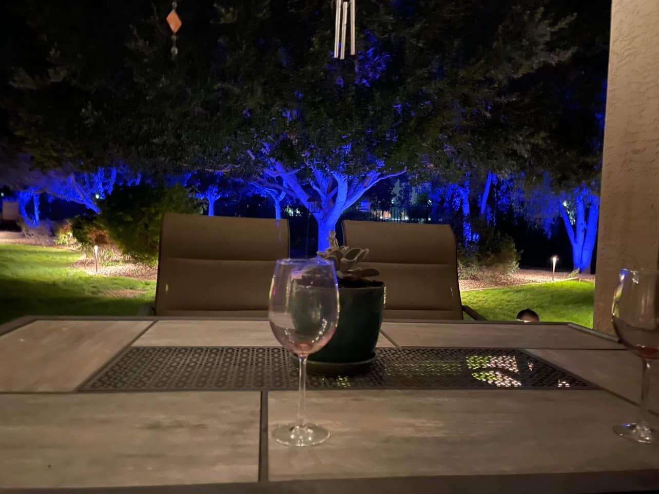 Patio table with wine glasses at night, overlooking blue-lit trees and outdoor chairs