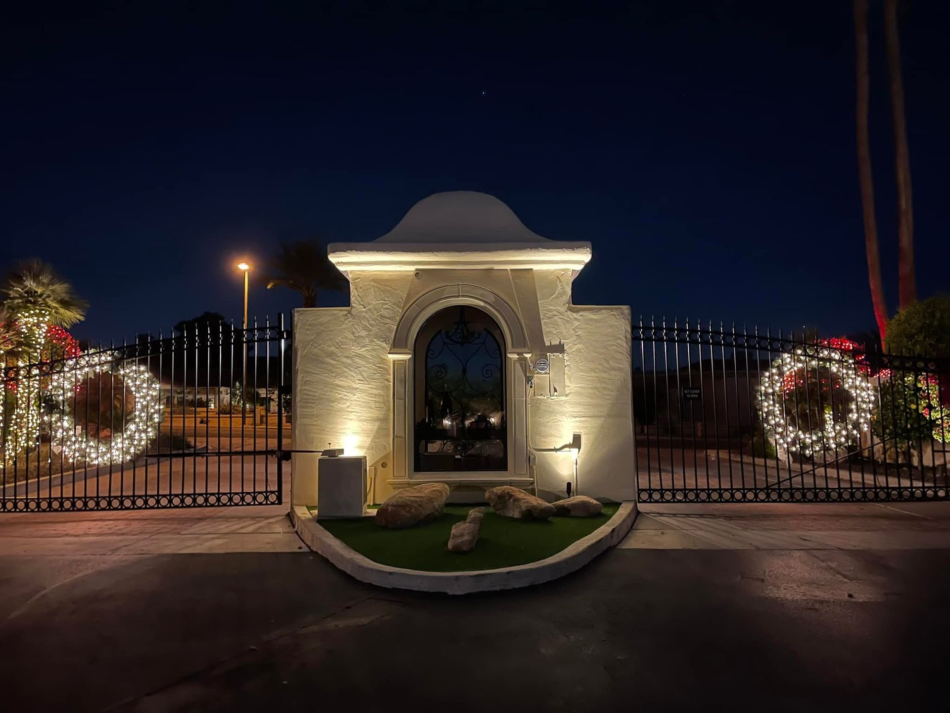 Illuminated stone entrance gate at night, flanked by decorative wreaths and a black iron fence.