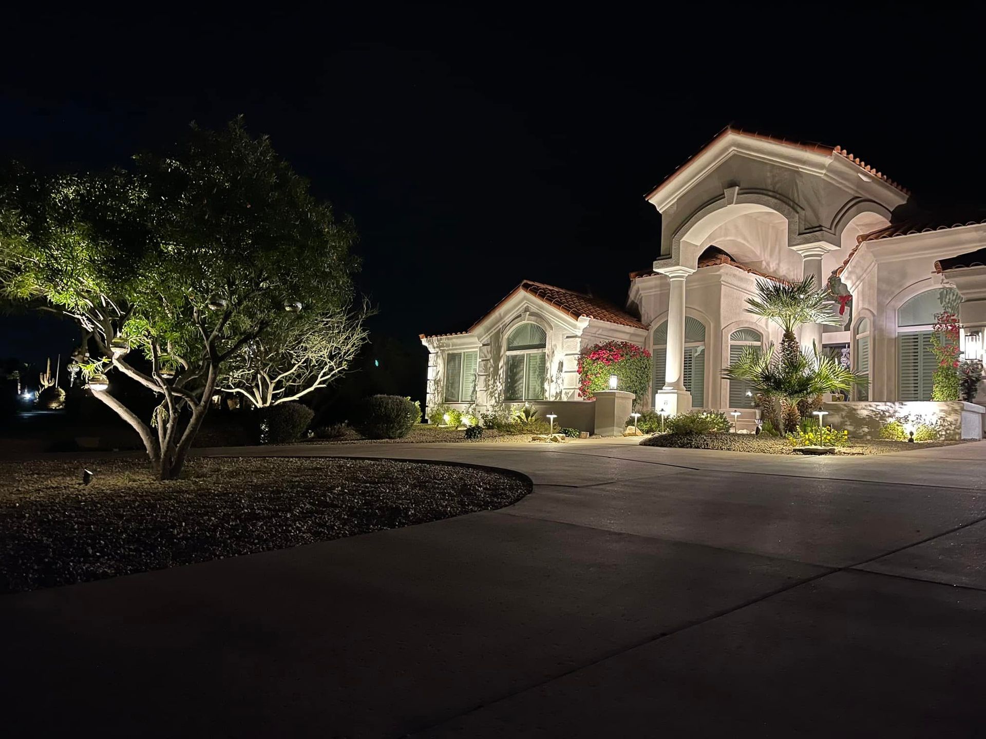 Nighttime view of a large, well-lit house with a landscaped driveway and trees