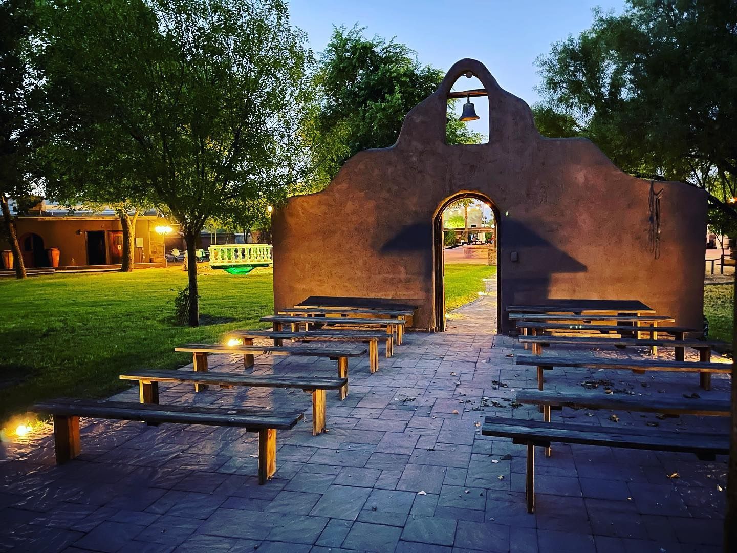 Stone courtyard with benches facing a small chapel entrance at dusk, lit by warm lights and surrounded by trees