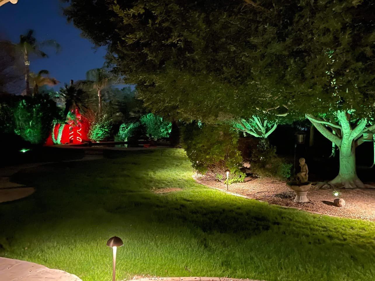 Nighttime garden path lit by green and red landscape lights, with trees and a grassy lawn