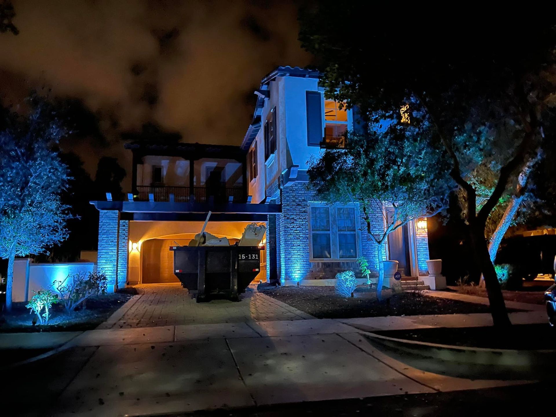 Modern house at night, lit with blue and orange lights under dark trees