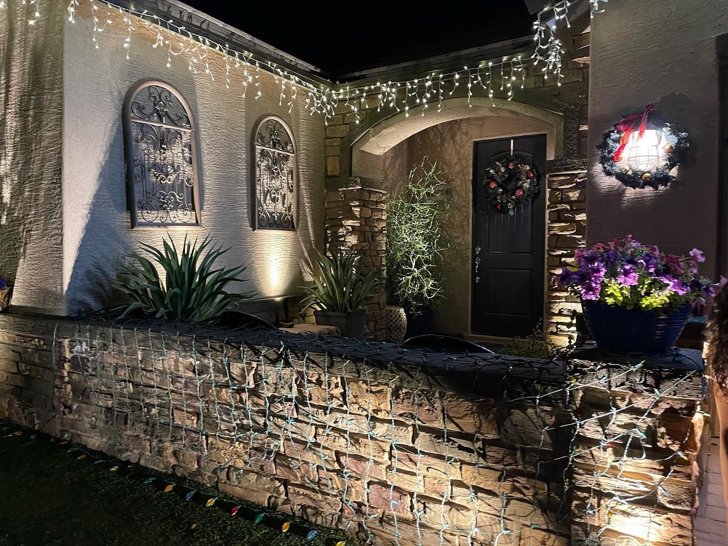 Nighttime brick courtyard with arched windows, warm lights, and a lit front door with flowers and plants