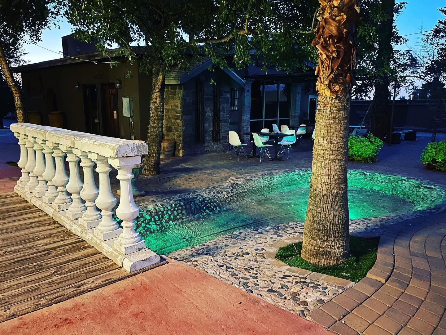 Tree-lined patio with white balustrade, outdoor chairs, and patterned paving beside a house at dusk