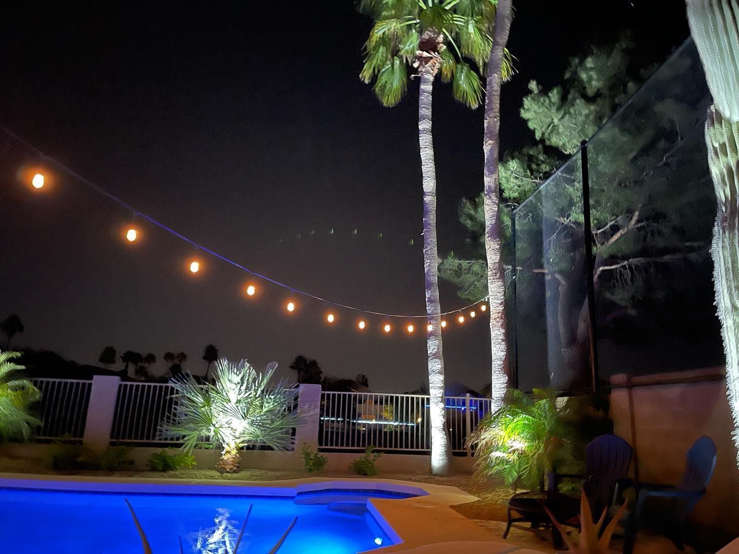 Nighttime poolside patio with string lights, palm trees, and glowing blue pool water