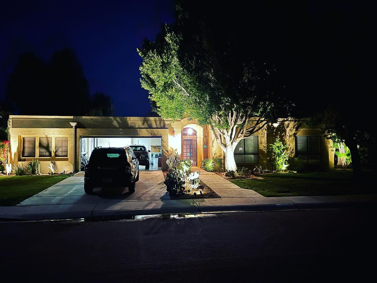 Nighttime view of a lit suburban house with a car in the driveway and trees in the yard.