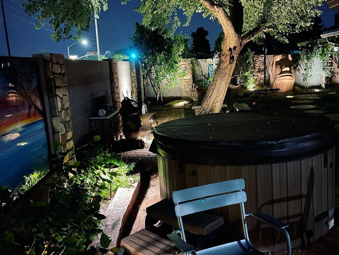 Backyard at night with a lit hot tub, stone wall, trees, and a white chair in the foreground