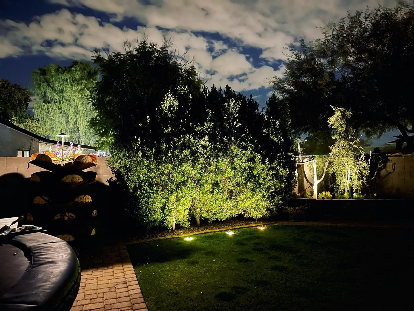 Backyard at night with lit trees, lawn, stone patio, and cloudy sky