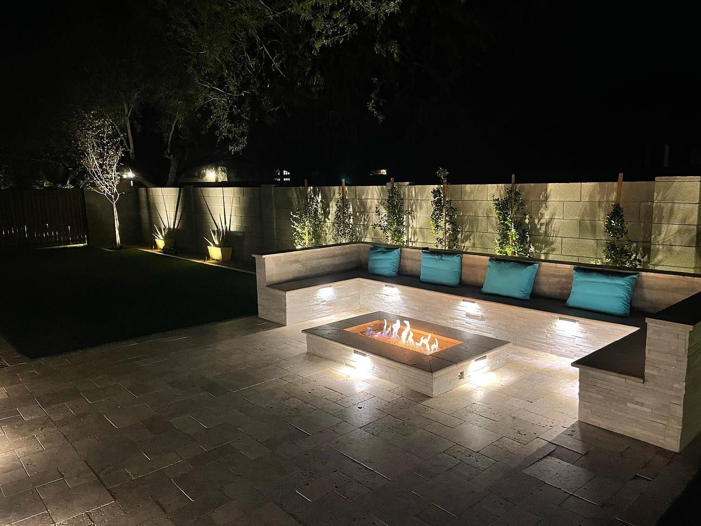 Outdoor patio at night with a built-in fire pit, sectional seating, and soft garden lighting