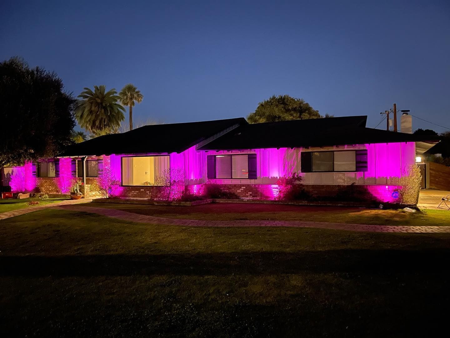 Single-story house lit with bright pink lights at dusk, with a dark lawn and trees in the background