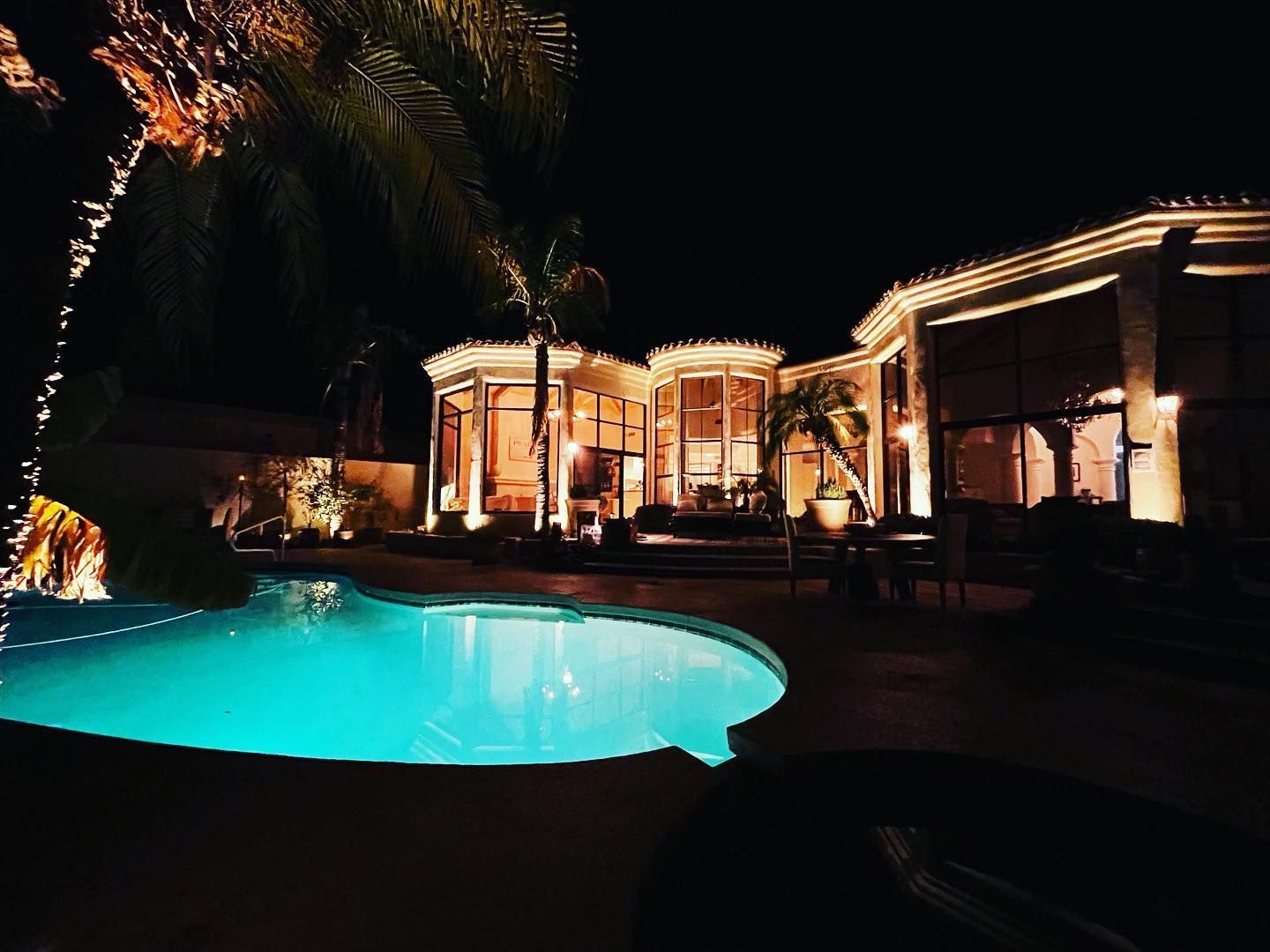 Nighttime patio with a glowing turquoise pool and warmly lit house exterior