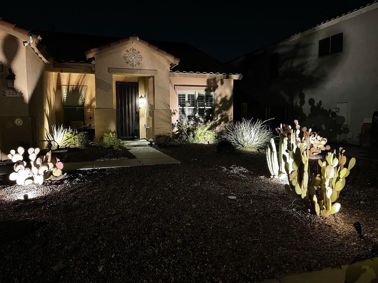 Nighttime view of a house entrance with lit landscaping and a dark gravel driveway.
