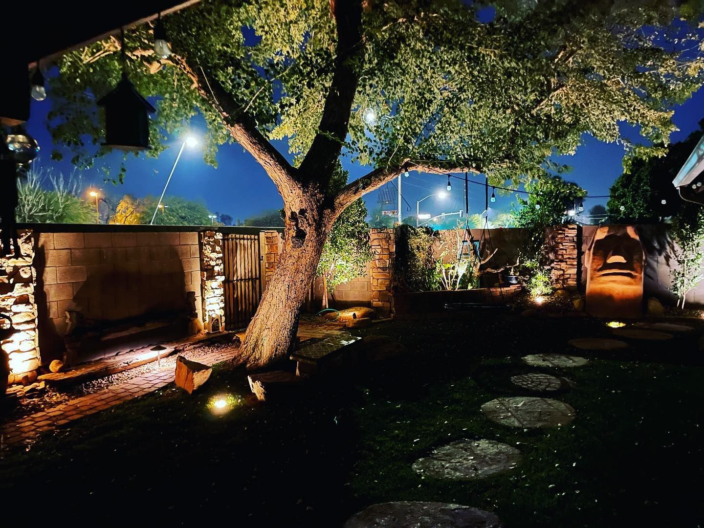 Illuminated backyard garden at night with a large tree, stone walkway, and brick walls
