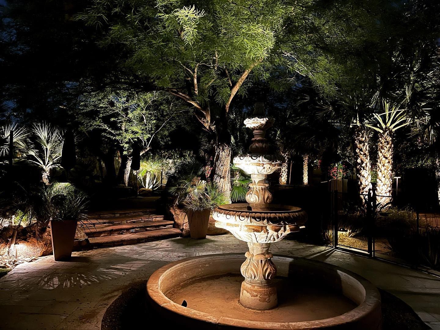 Stone fountain in a dimly lit garden courtyard with trees and potted plants.