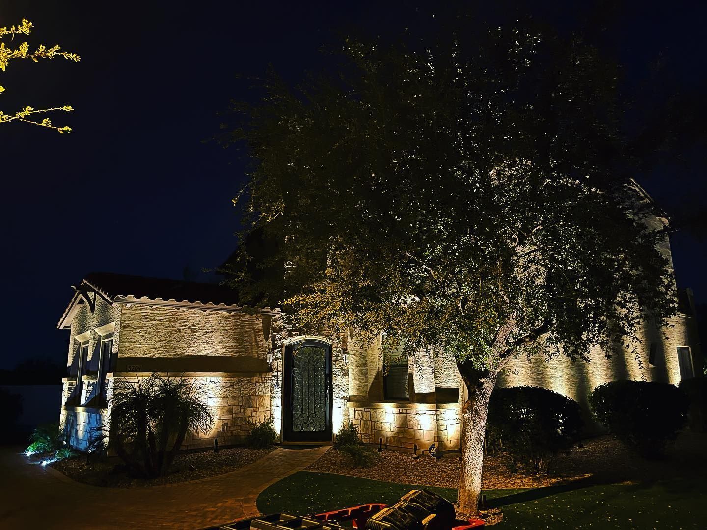 Night-lit stone house and tree in a garden, warmly illuminated against a dark sky.