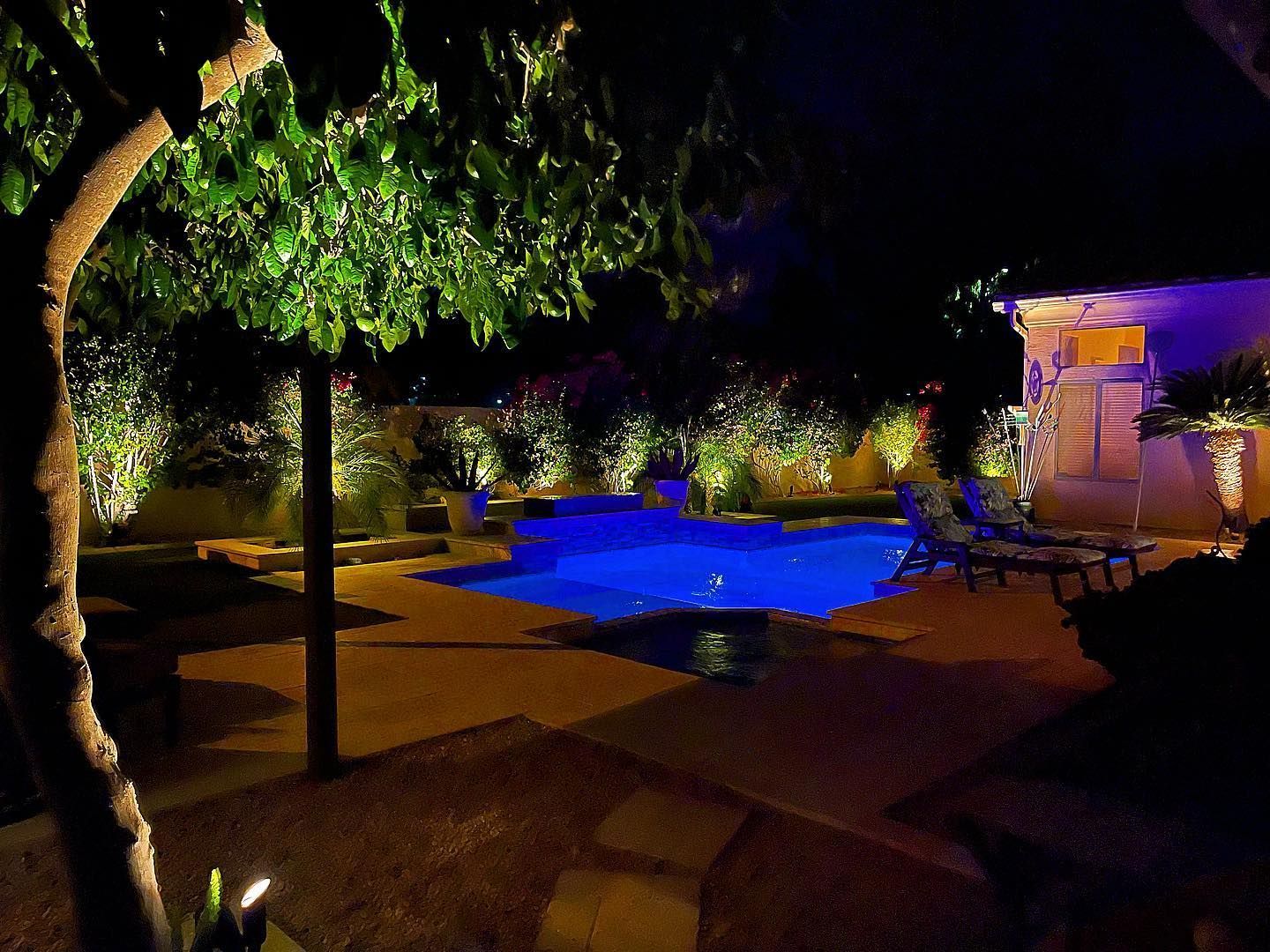 Nighttime poolside patio with blue-lit swimming pool, trees, and a lit house in the background