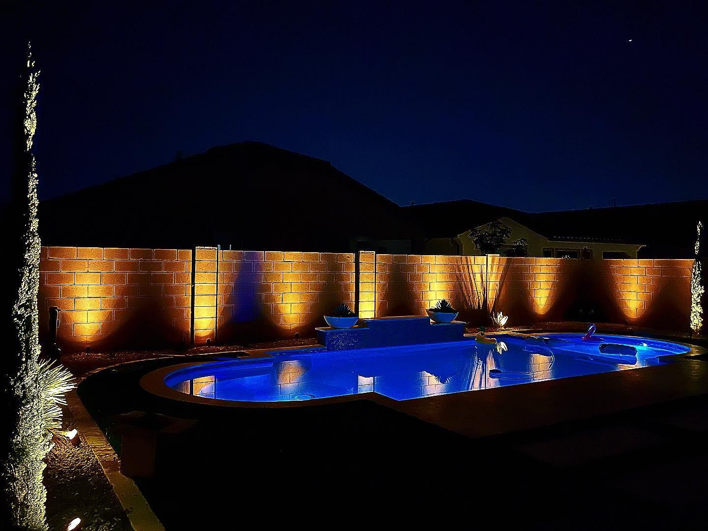Nighttime pool with blue water, warm-lit stone wall, and dark mountains under a clear sky