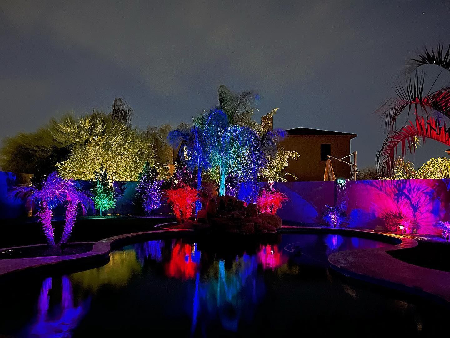 Colorfully lit garden and pool at night with blue, red, and purple reflections on the water