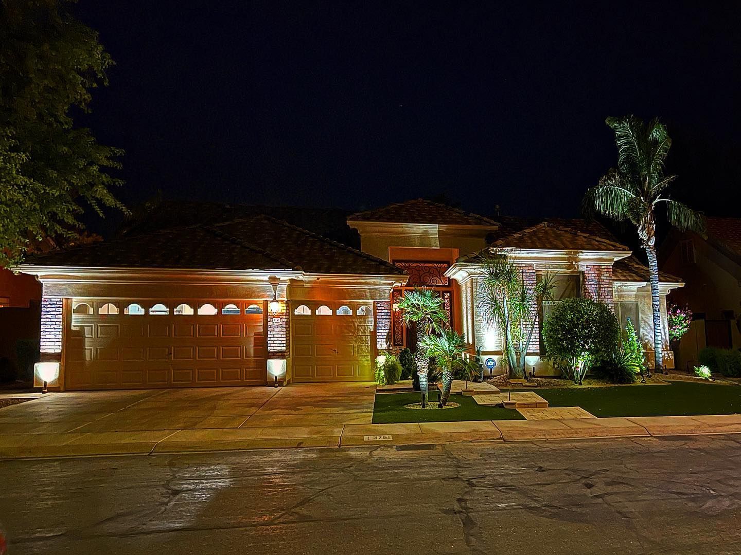 Nighttime view of a lit suburban house with garage, palm trees, and a wet driveway