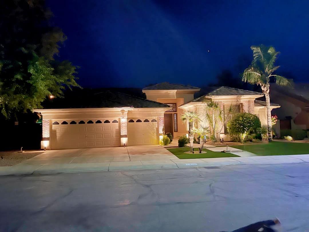 Suburban house at night with garage lights on, palm trees, and a driveway under a dark blue sky