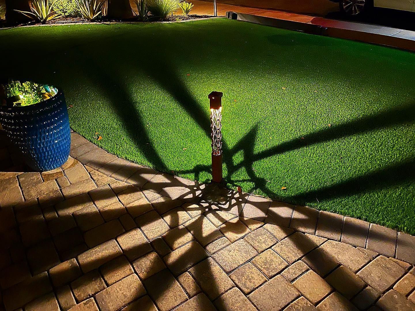 Sunlit brick patio beside a green lawn, with a blue planter and a small garden statue casting long shadows.