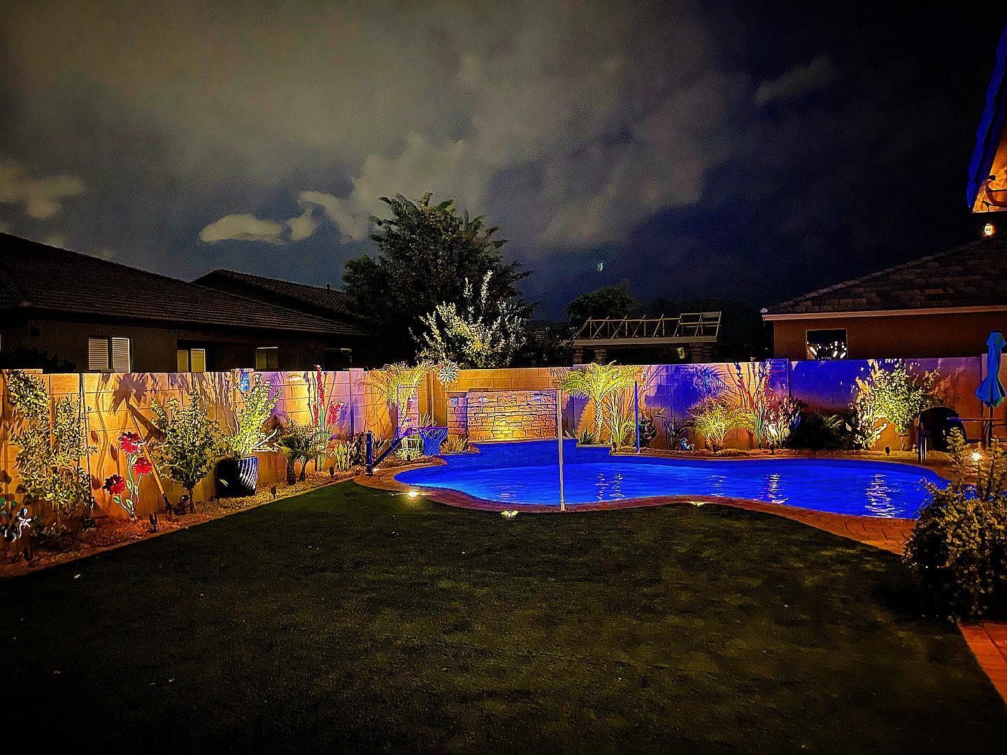 Nighttime backyard with a lit swimming pool, colorful garden lights, and a dark lawn under cloudy sky