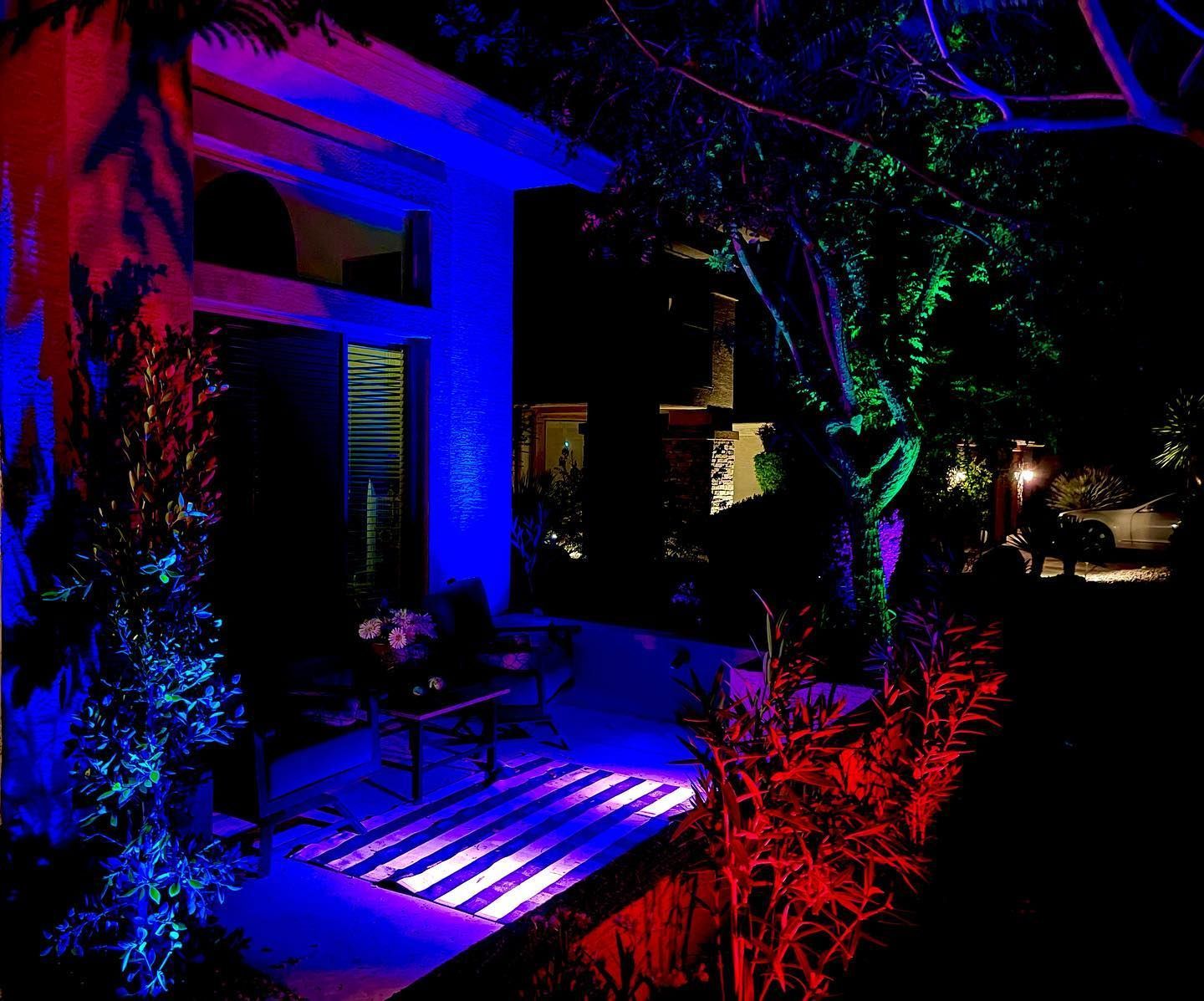 Night-lit house entrance with blue and red lights illuminating the porch and walkway, surrounded by trees and shrubs