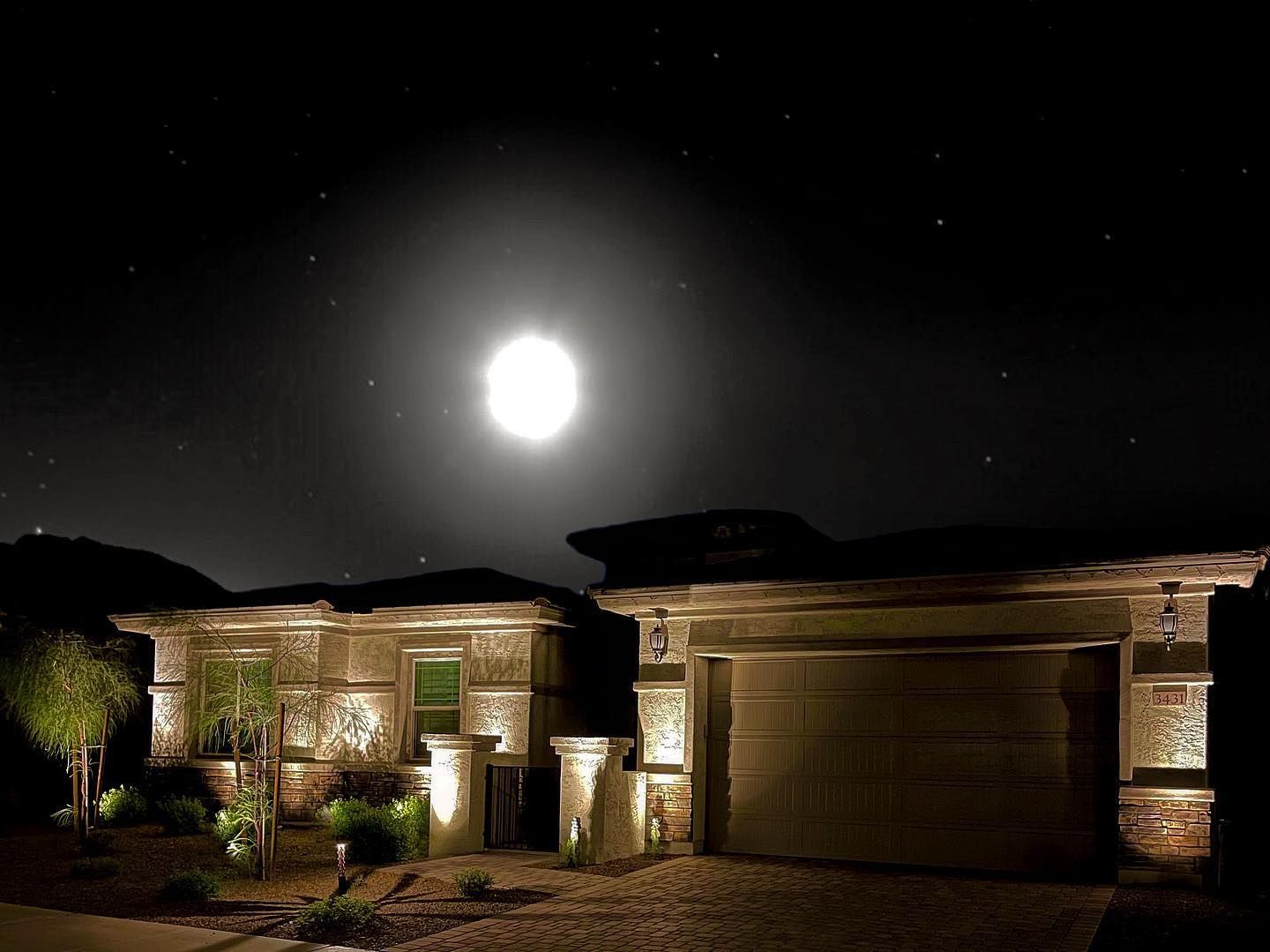 Nighttime view of a lit brick house with a garage under a bright moon and starry sky