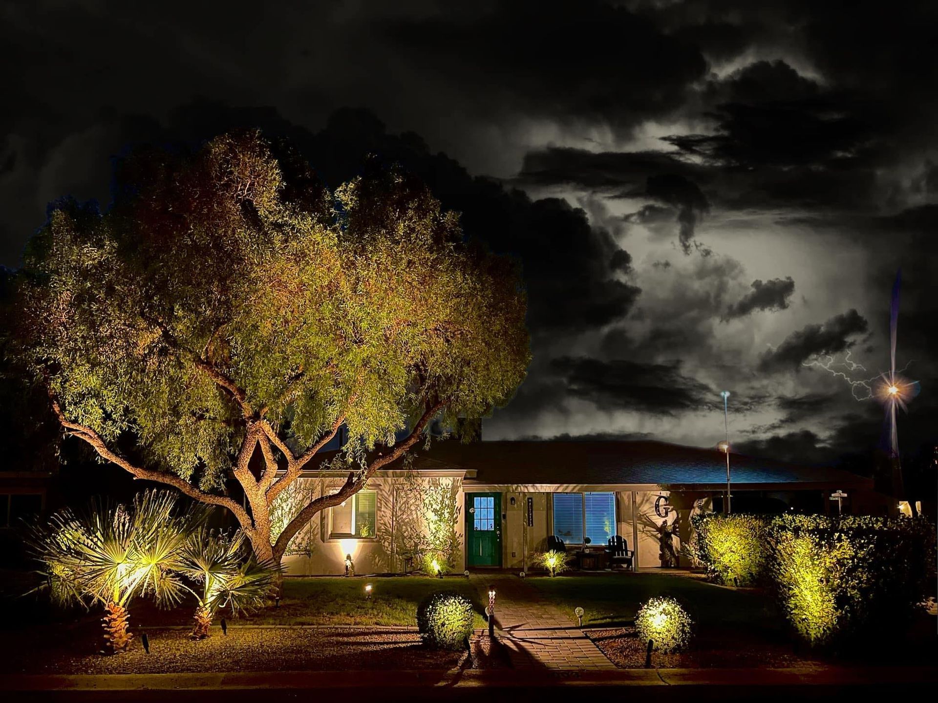 A lit house and large tree at night under dark, cloudy skies with a glowing yard and streetlight.