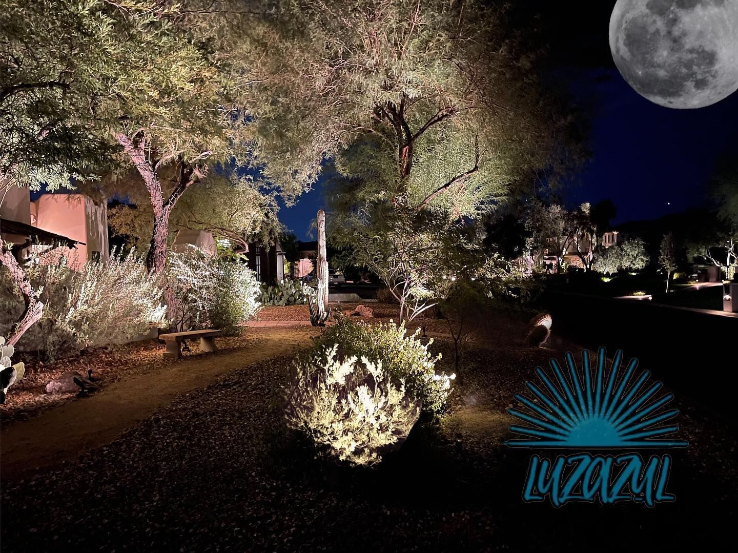Moonlit garden path with lit trees and shrubs, and a teal logo in the lower right corner