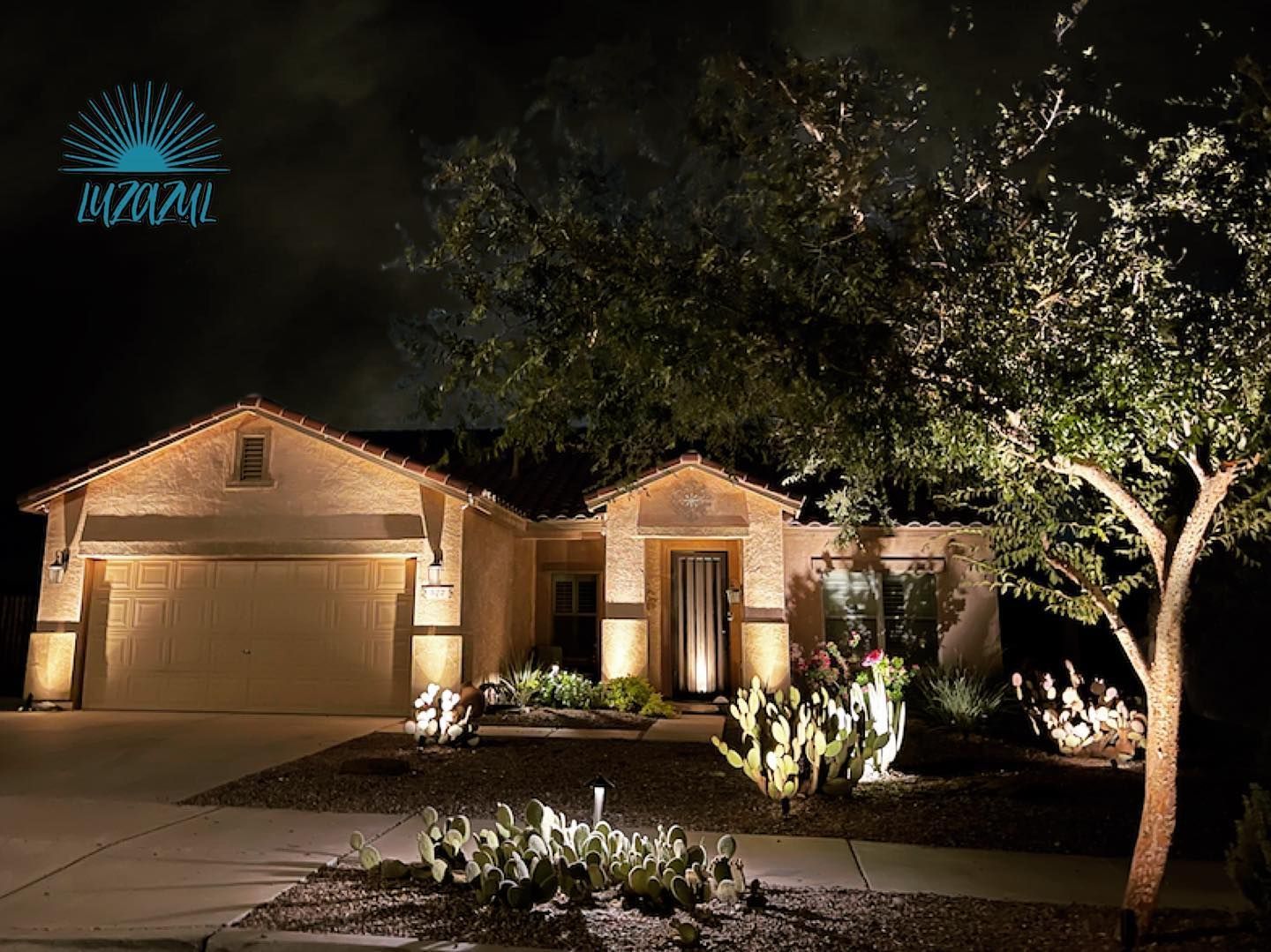 Suburban house at night with warm exterior lights and a large tree in the front yard