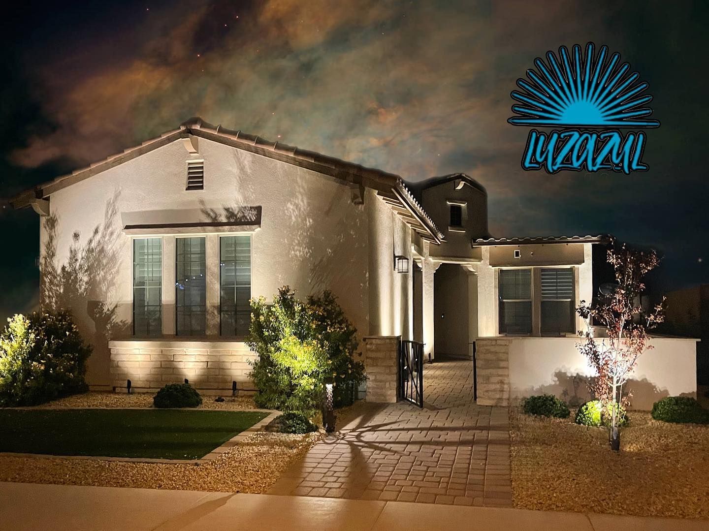 Well-lit white stucco house at night with blue neon sign in the sky, landscaped front yard, and stone walkway