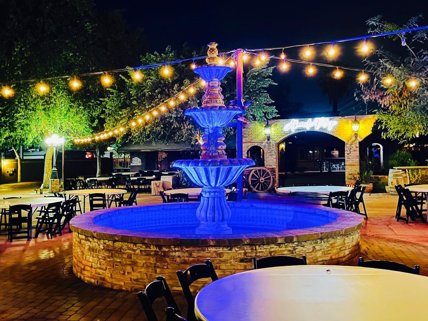Illuminated fountain in an outdoor patio at night with string lights and tables around it