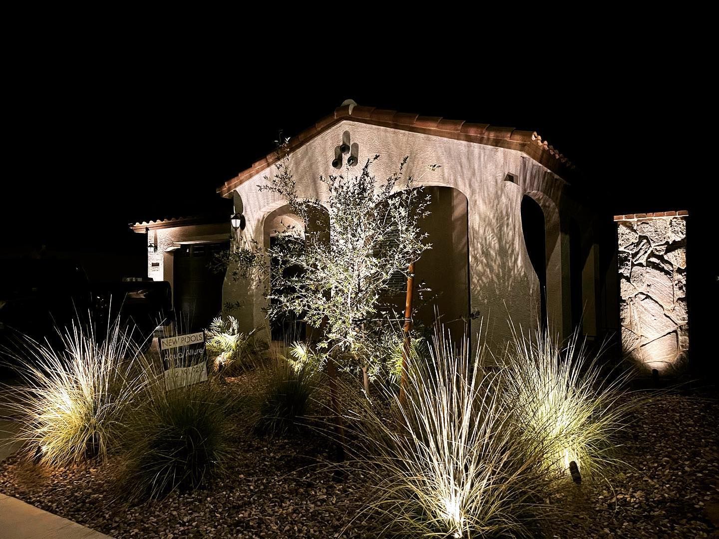 Well-lit house at night with illuminated landscaping and ornamental grasses in front