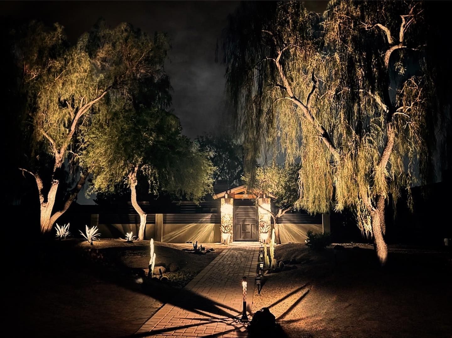 Nighttime garden scene with trees, a lit pathway, and a small gate or shrine in the center.