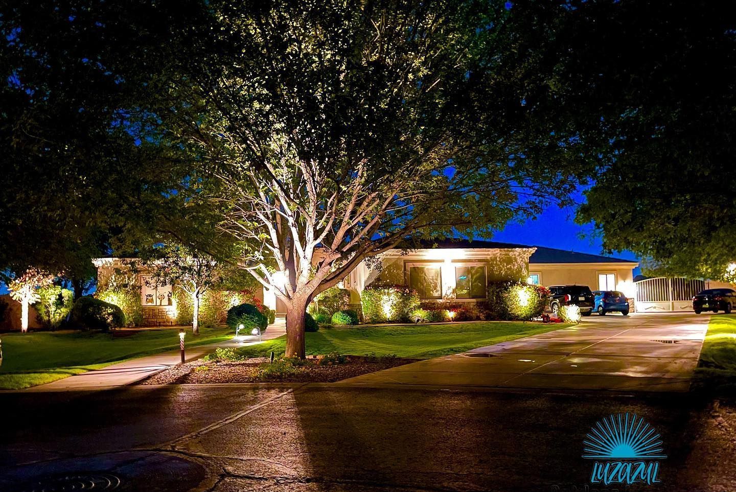 Nighttime view of a lit house and large tree in a suburban driveway, with blue sky and wet pavement