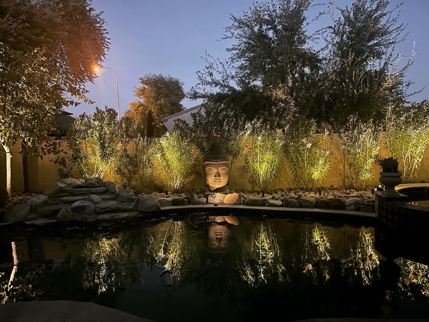 Twilight garden with a reflective pond, lit trees, and glowing lights along the far bank